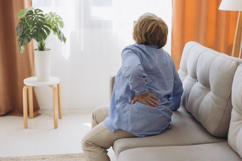 Unhappy old woman touching back, feeling pain, sitting on couch in living room, unhealthy middle aged senior female suffering from backache, spinal
